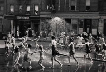 Children in swimsuits hold hands in a circle around a spray of water from a fire hydrant on a city street, with several adults and buildings in the background. The scene appears joyful and lively.