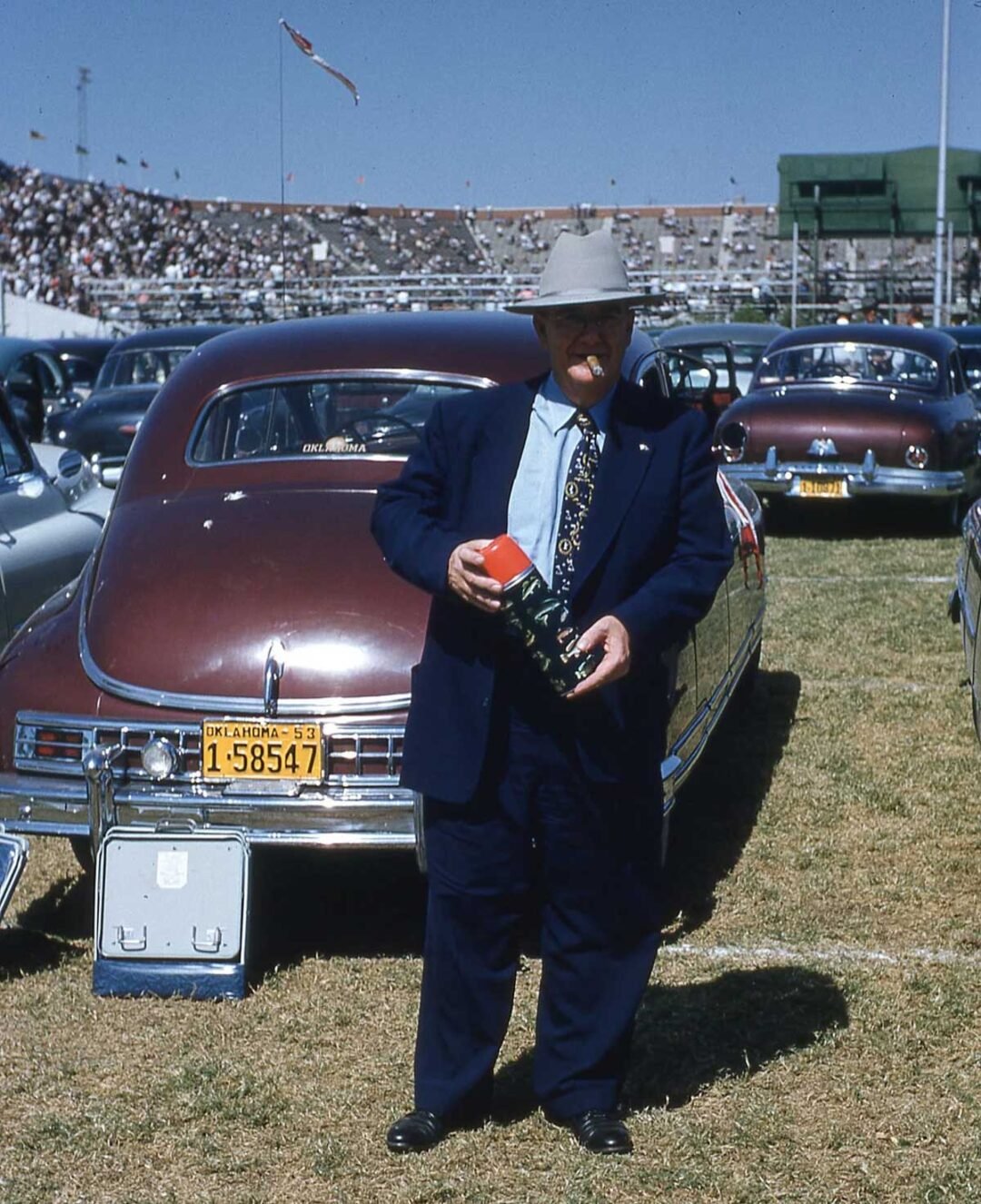 An older man in a suit and hat stands on grass behind a vintage car with an Oklahoma license plate, holding a camera. Stadium seats and a large crowd are visible in the background under a clear sky.