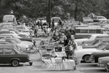 Black and white photo of people tailgating between rows of parked cars, with folding chairs, tables, picnic food, and American flags, surrounded by trees in the background.