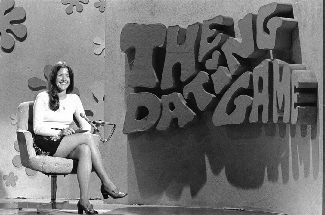 A woman sits smiling on a swivel chair next to a large, playful sign reading "The Dating Game" on a vintage television set. The scene is in black and white.