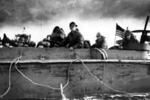 A group of World War II soldiers in helmets and gear sit inside a military landing craft on water, with one looking over the side; an American flag is visible at the back under a cloudy sky.