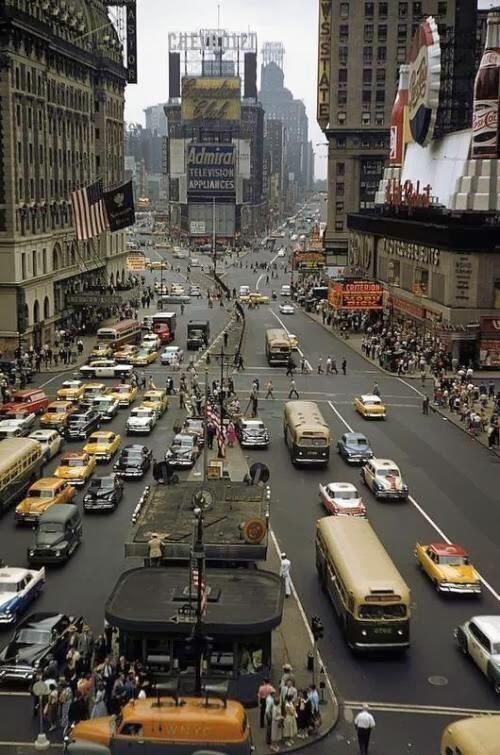 A busy city intersection filled with cars, taxis, and buses, surrounded by tall buildings with colorful advertisements; many pedestrians cross the street. The image has a vintage, mid-20th-century feel.