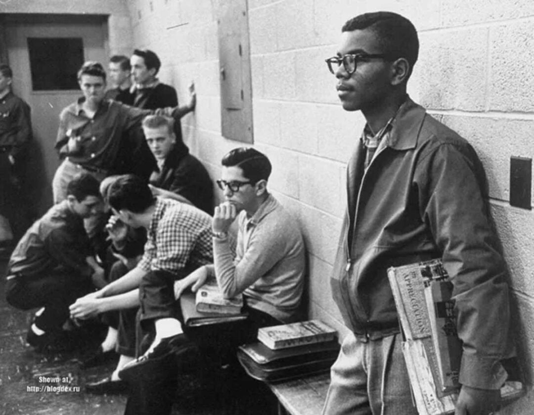 A young Black student stands alone holding books, while a group of white students sit and talk together on benches in a school hallway, highlighting a scene of racial segregation.