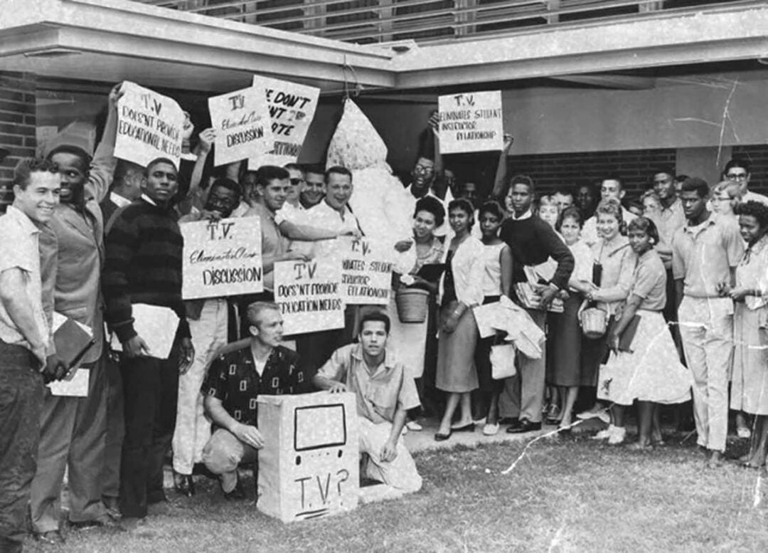 A large group of students gathers outside a building, holding protest signs with phrases like "TV Doesn't Provide Education," "TV Causes Division," and "TV Destroys Student Involvement." Some hold a TV-shaped prop.
