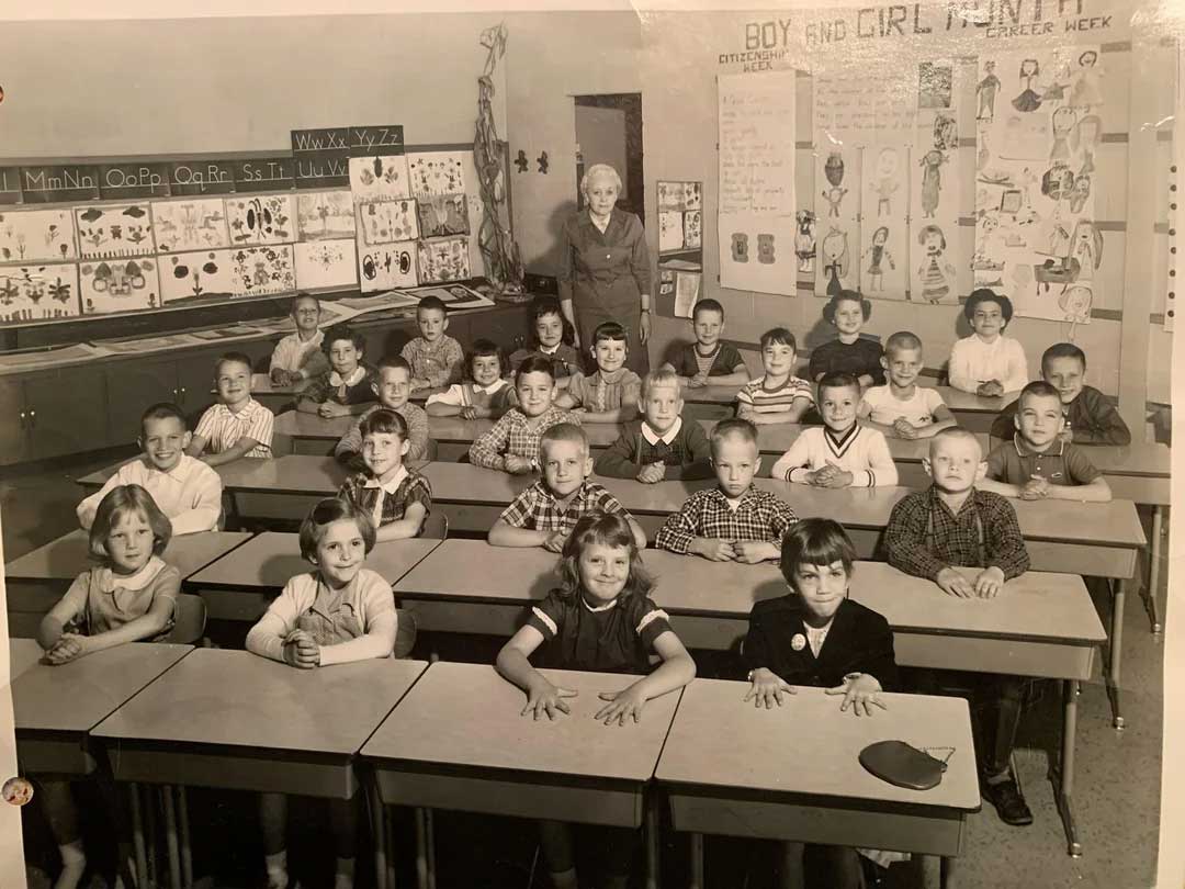 A vintage black and white photo of a classroom with young children seated at desks in rows and a female teacher standing at the back. Educational posters and artwork decorate the classroom walls.