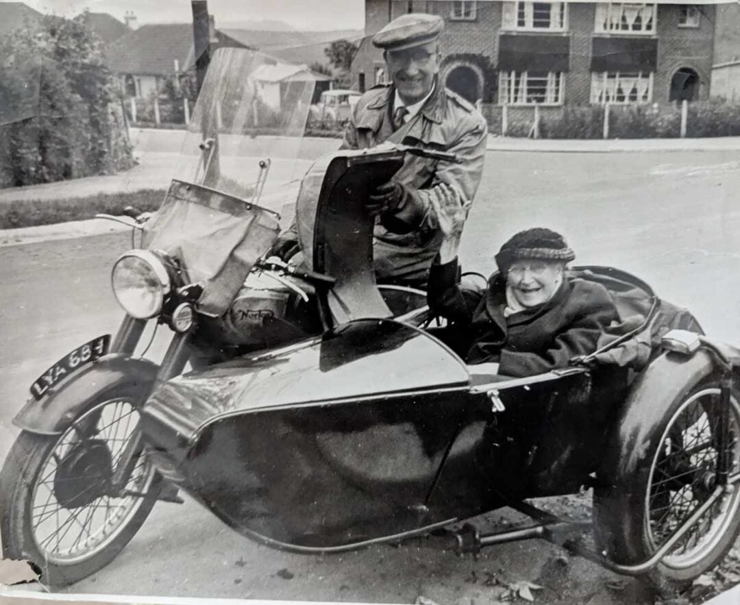 A smiling man sits in the driver’s seat of a vintage motorcycle with a sidecar, while an elderly woman sits happily in the sidecar. They are outdoors in a residential neighborhood.