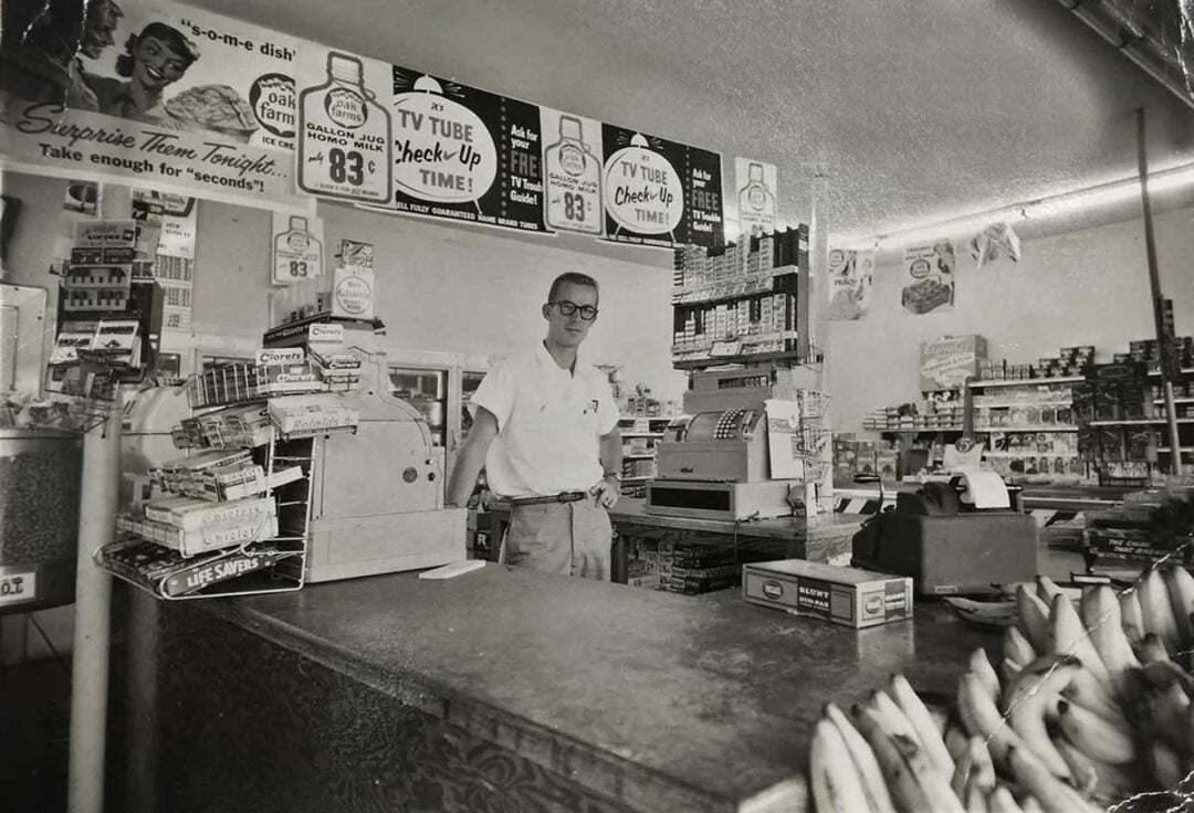 A man wearing glasses and a white shirt stands behind the counter of a vintage grocery store filled with products, cash registers, and signs advertising TV tube check-ups and soup. Bananas and snacks are visible in the foreground.
