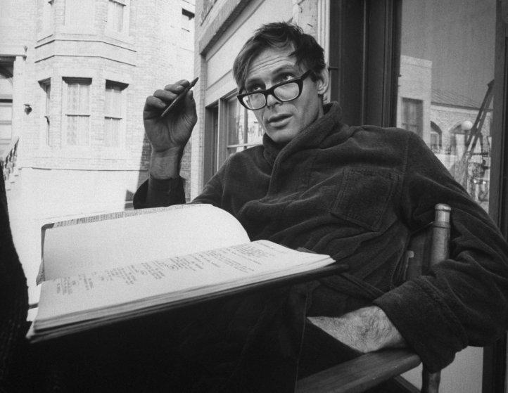 A man wearing glasses and a robe sits in a chair outdoors, holding a pen and looking up thoughtfully. An open book or script with handwritten notes rests on his lap. Brick buildings are visible in the background.