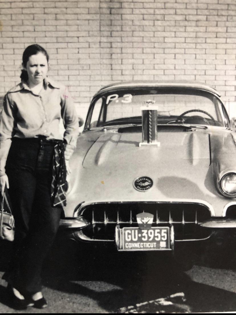 A woman stands next to a vintage car with a Connecticut license plate and a large trophy displayed on the hood. The background is a brick wall. The photo is black and white.