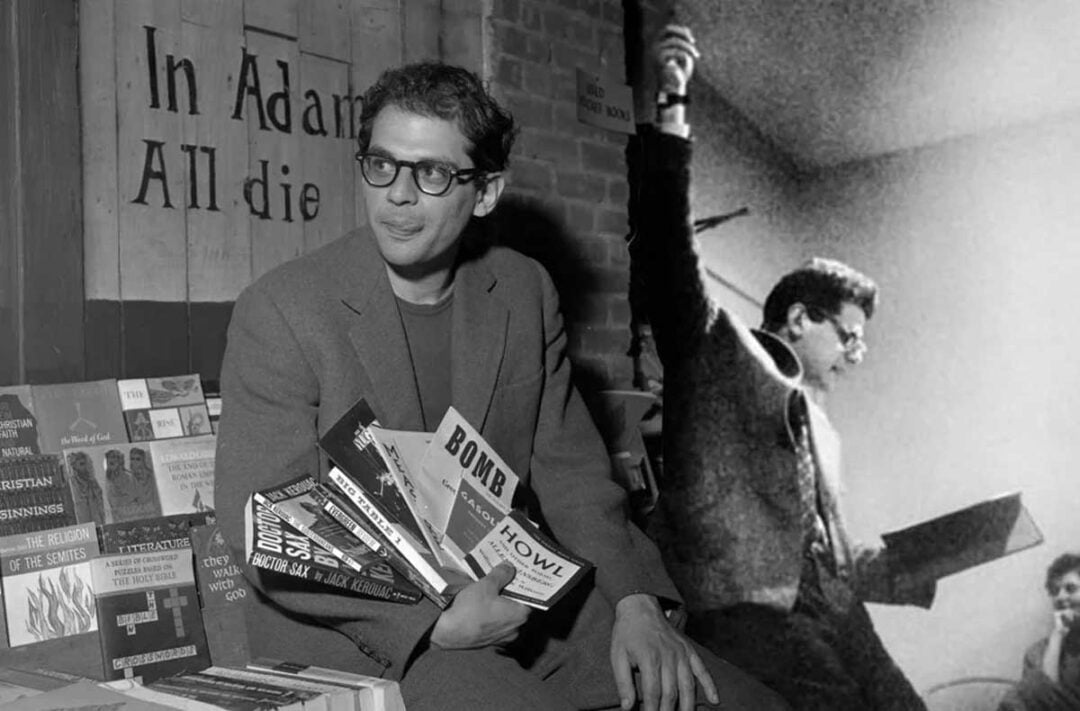 A man in glasses and a suit sits holding several books in one hand, surrounded by books on display. Behind him, there’s a large black-and-white image of the same man passionately reading aloud to an audience.