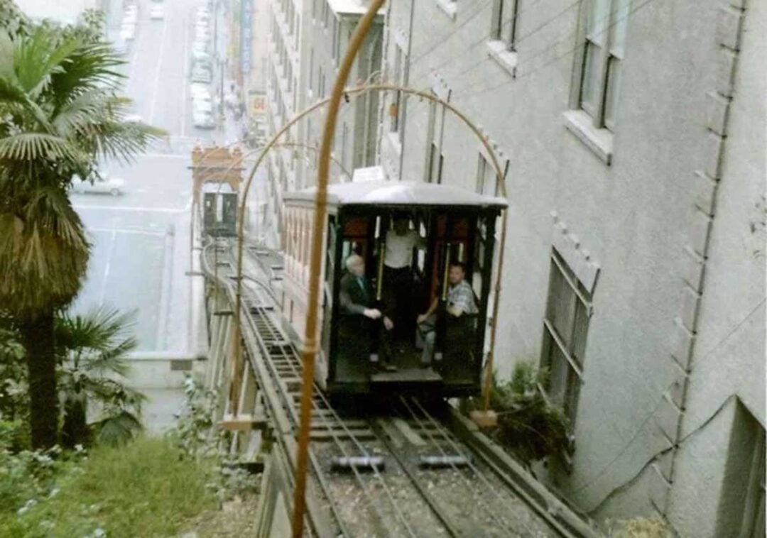 A historic funicular railway car with several passengers ascends a steep hill between two buildings, with trees and a city street visible in the background.