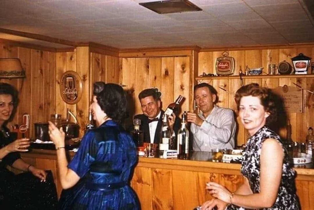 Four adults in 1950s-style clothing gather around a wood-paneled home bar; two men stand behind the bar mixing drinks, while two women sit in front, smiling and holding cocktails.