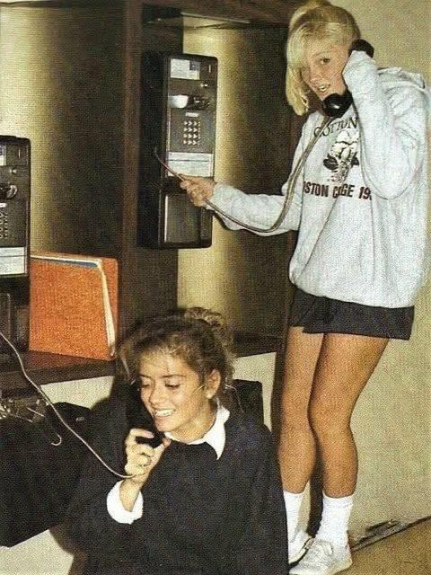 Two young women use corded payphones; one is standing, wearing a hoodie and shorts, while the other is crouched, holding a phone with a smile. Both appear to be in a casual, indoor setting.
