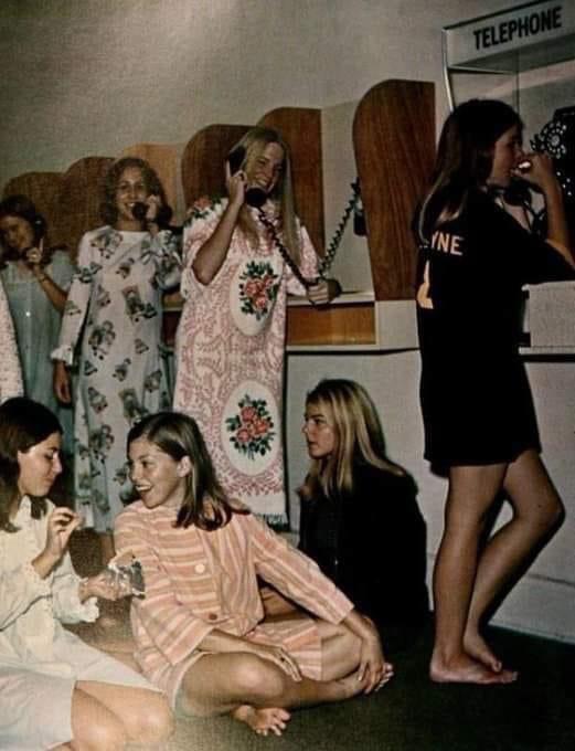 Six teenage girls in pajamas and nightgowns are gathered in a room with multiple payphones. Some are talking on the phones while others sit and chat on the floor, creating a relaxed, nostalgic sleepover atmosphere.