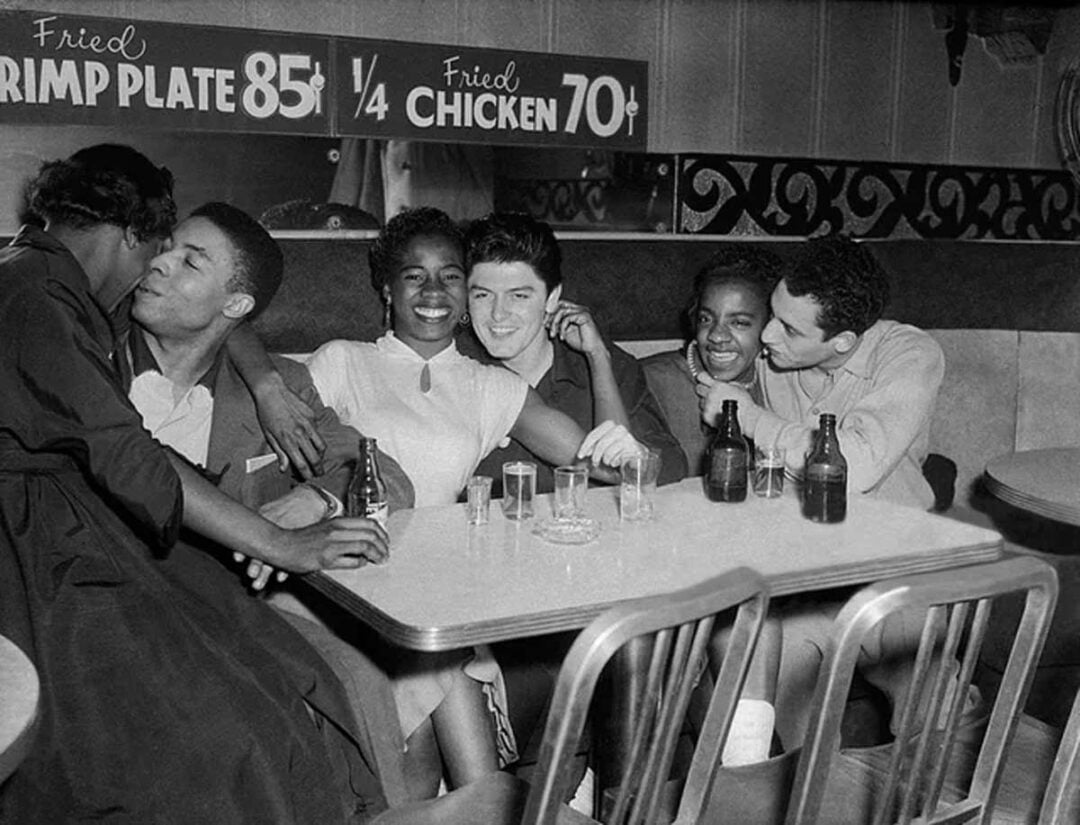 Five young adults, three women and two men, sit closely together at a restaurant table, smiling and laughing. Sign behind them advertises fried shrimp plate for 85 cents and quarter fried chicken for 70 cents.