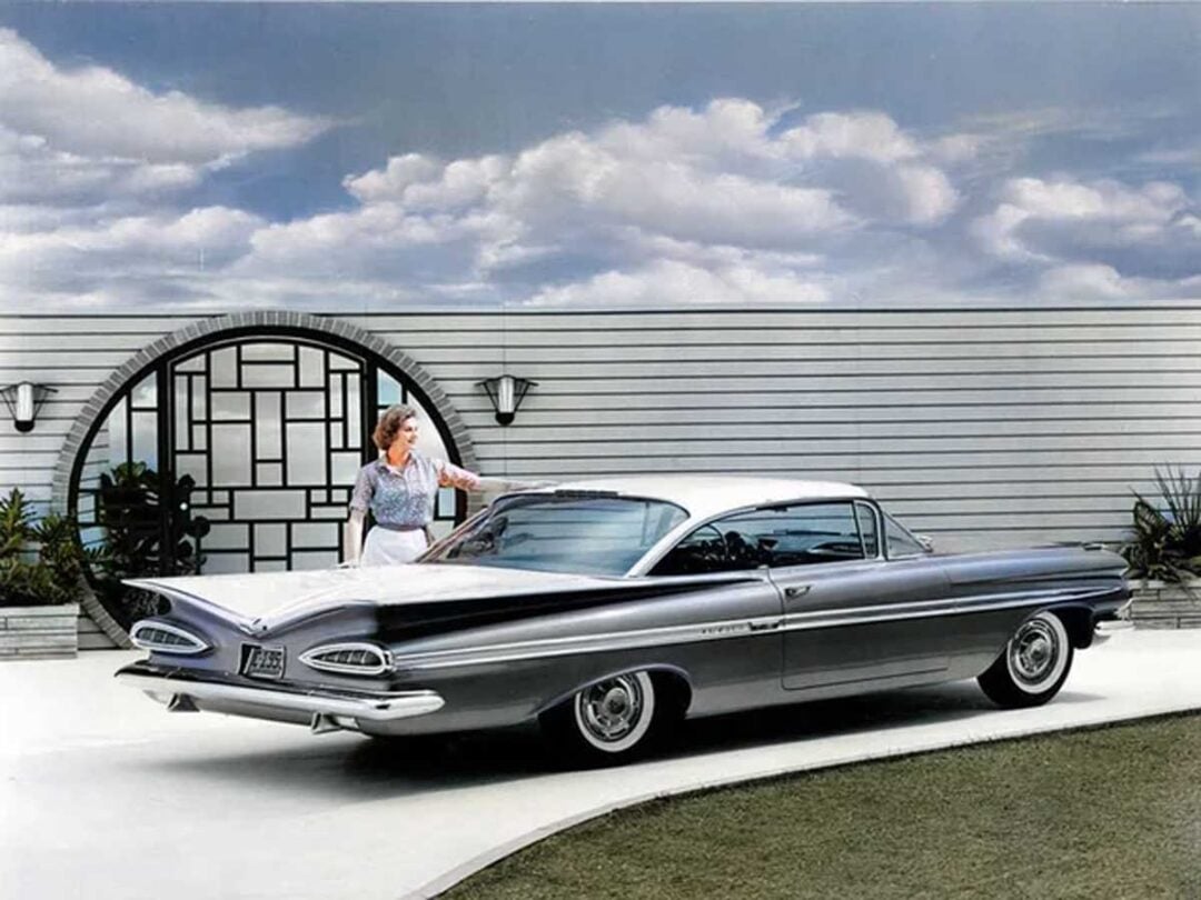 A woman stands beside a classic silver and white car with large tail fins, parked on a driveway in front of a modern mid-century house with a circular gate and striped walls under a partly cloudy sky.