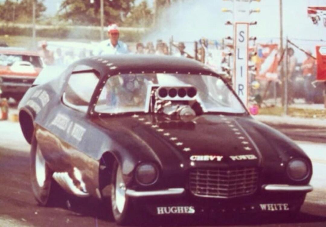 A vintage drag racing car with “Hughes” and “White” written on the front performs on a racetrack. Smoke rises from the rear tires as spectators and officials watch in the background.