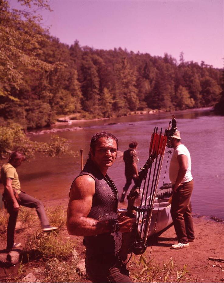 A muscular man holding a bow and arrows stands by a riverbank, looking at the camera. Two other men prepare a canoe behind him, surrounded by trees and greenery under a clear sky.