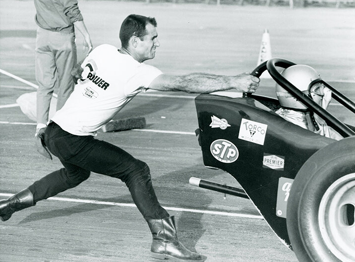 A man in a t-shirt and boots pushes a dragster race car from behind on a paved track; the driver is seated and wearing a helmet. The car displays various sponsor decals.
