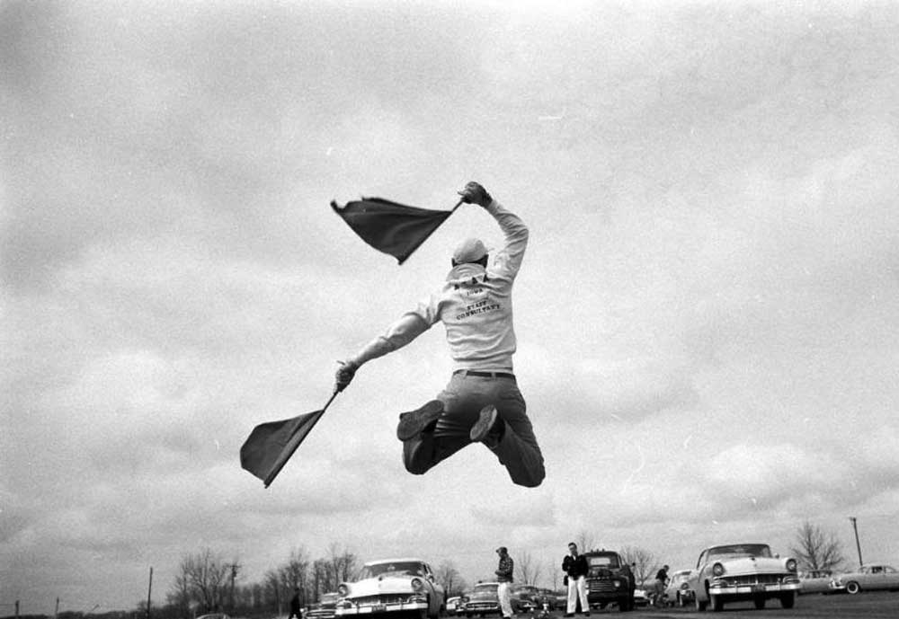 A person leaps into the air holding two flags, with vintage cars and people in the background under a cloudy sky. The scene appears energetic, with an open road and bare trees in the distance.