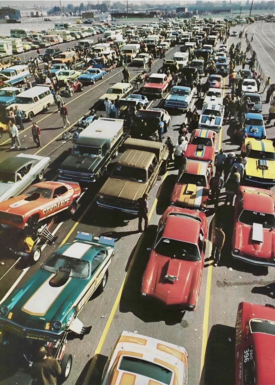 An aerial view of a parking lot filled with rows of colorful vintage race cars, drivers, and people gathered around, preparing for a motorsport event under a clear sky.