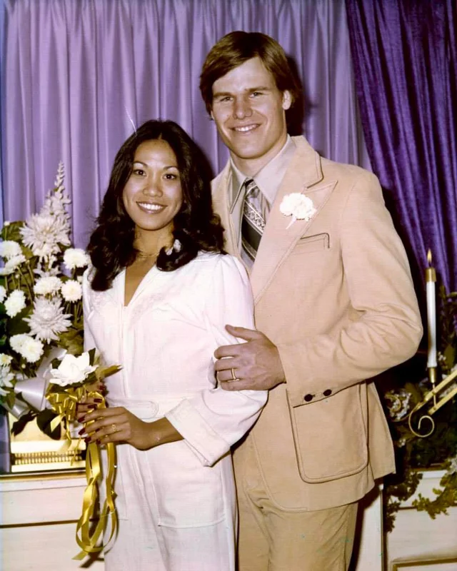 A smiling couple poses in front of purple curtains; the woman holds a bouquet with yellow ribbons and wears a white outfit, while the man stands beside her in a tan suit with a white flower on his lapel.