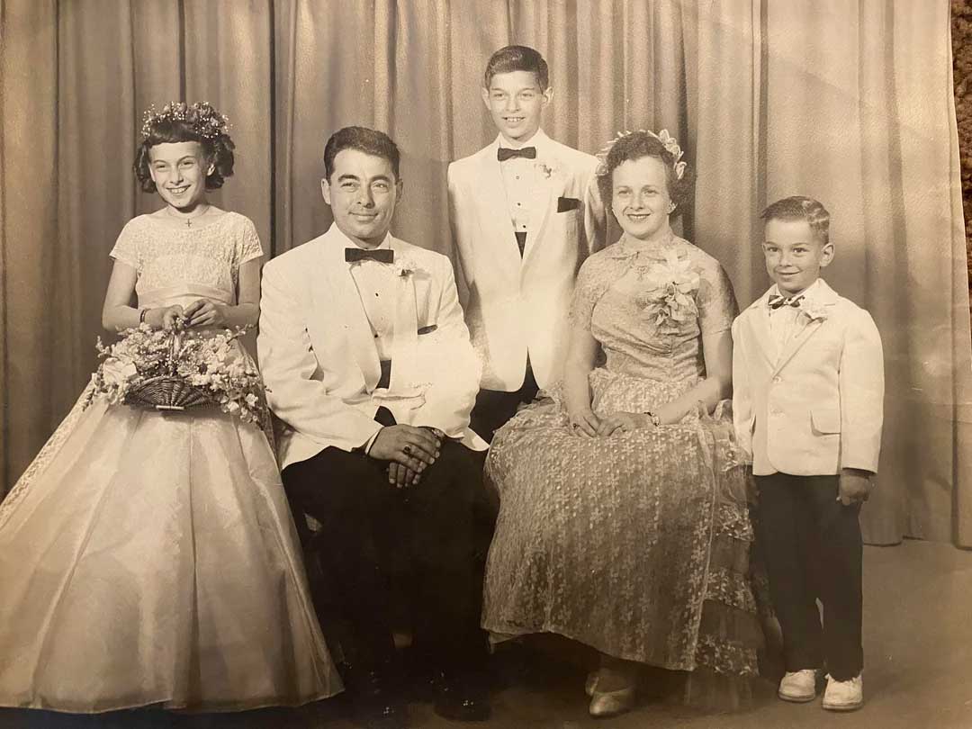 A vintage black-and-white photo of a family dressed formally. Two adults sit in the center, flanked by three children—two boys in suits and a girl in a dress with a flower crown—posing in front of a curtain backdrop.