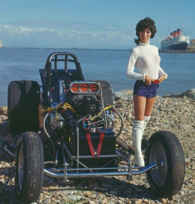 A woman in a white turtleneck, blue shorts, and white boots stands on a rocky shore beside a drag racing car, with water and a large ship in the background.
