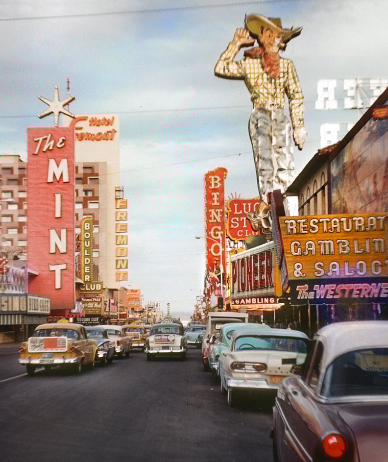 Colorful vintage photo of a busy Las Vegas street lined with neon signs, classic 1950s cars, and a large cowboy sign above the Pioneer Club. Crowds and casino advertisements fill the lively scene.
