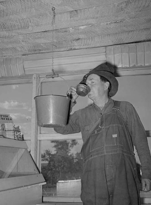A man in overalls and a hat drinks from a metal cup, pouring water from a large bucket suspended from the ceiling by a chain in a rustic indoor setting with windows and visible trees outside.