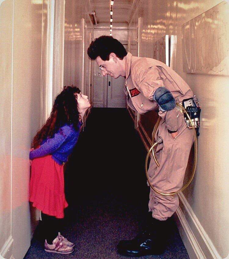 A man in a Ghostbusters uniform leans forward to face a young girl standing in a hallway. The girl, with long curly hair and a red skirt, looks up at him with her hands behind her back.