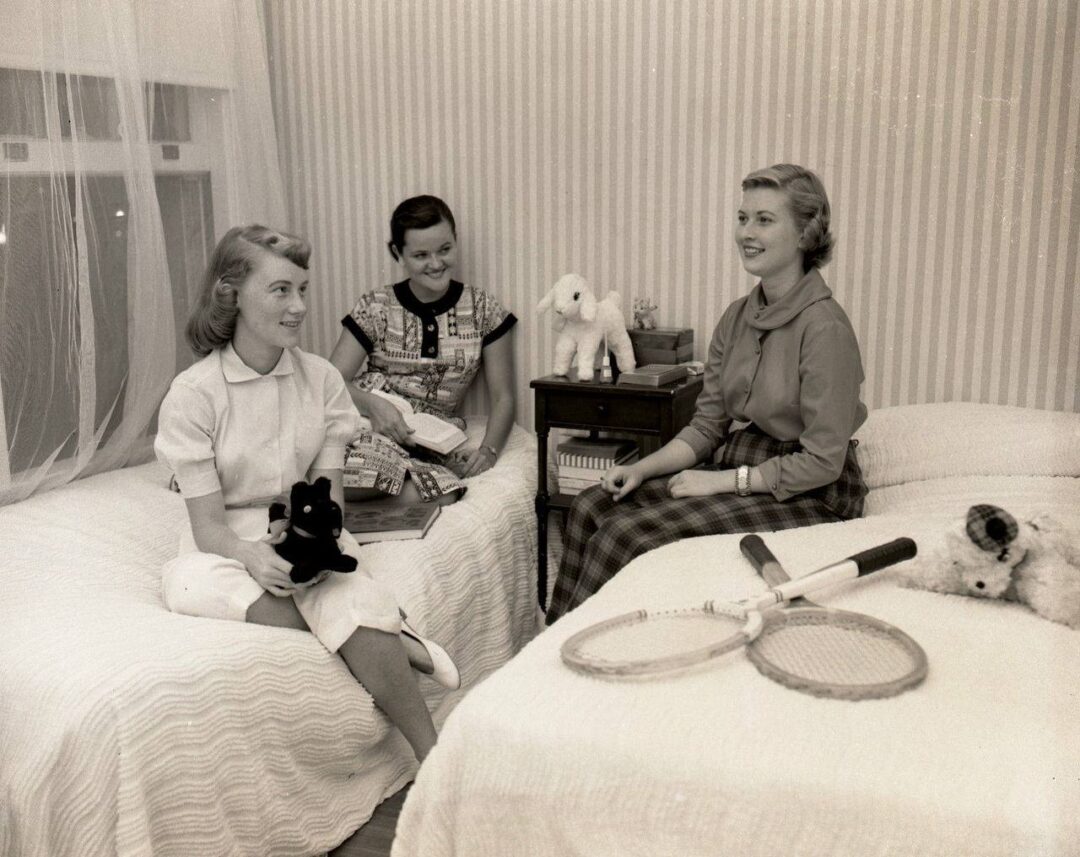 Three girls sit and chat in a neatly arranged bedroom with twin beds, stuffed animals, and tennis racquets. They appear relaxed and happy, dressed in 1950s-style clothing. A nightstand with a lamp and toys is between the beds.