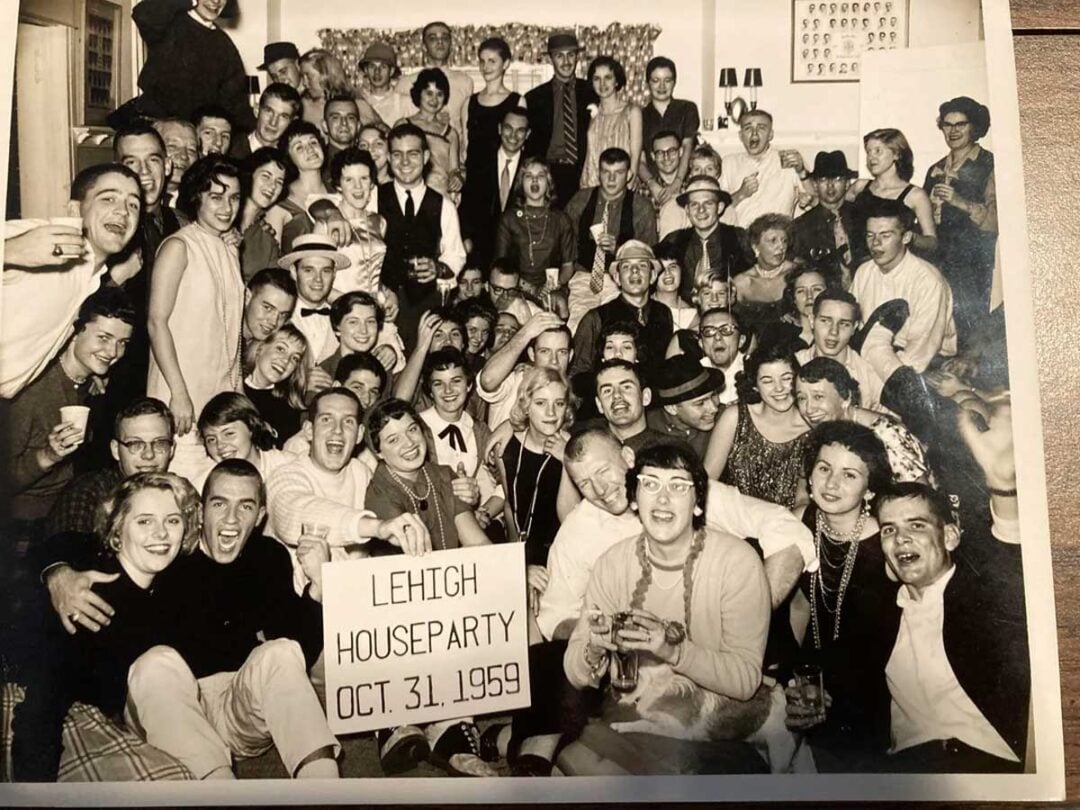 Large group of young adults in costume crowd together, smiling and celebrating at a house party. A sign in front reads “Lehigh Houseparty Oct. 31, 1959.” The atmosphere is festive and cheerful.