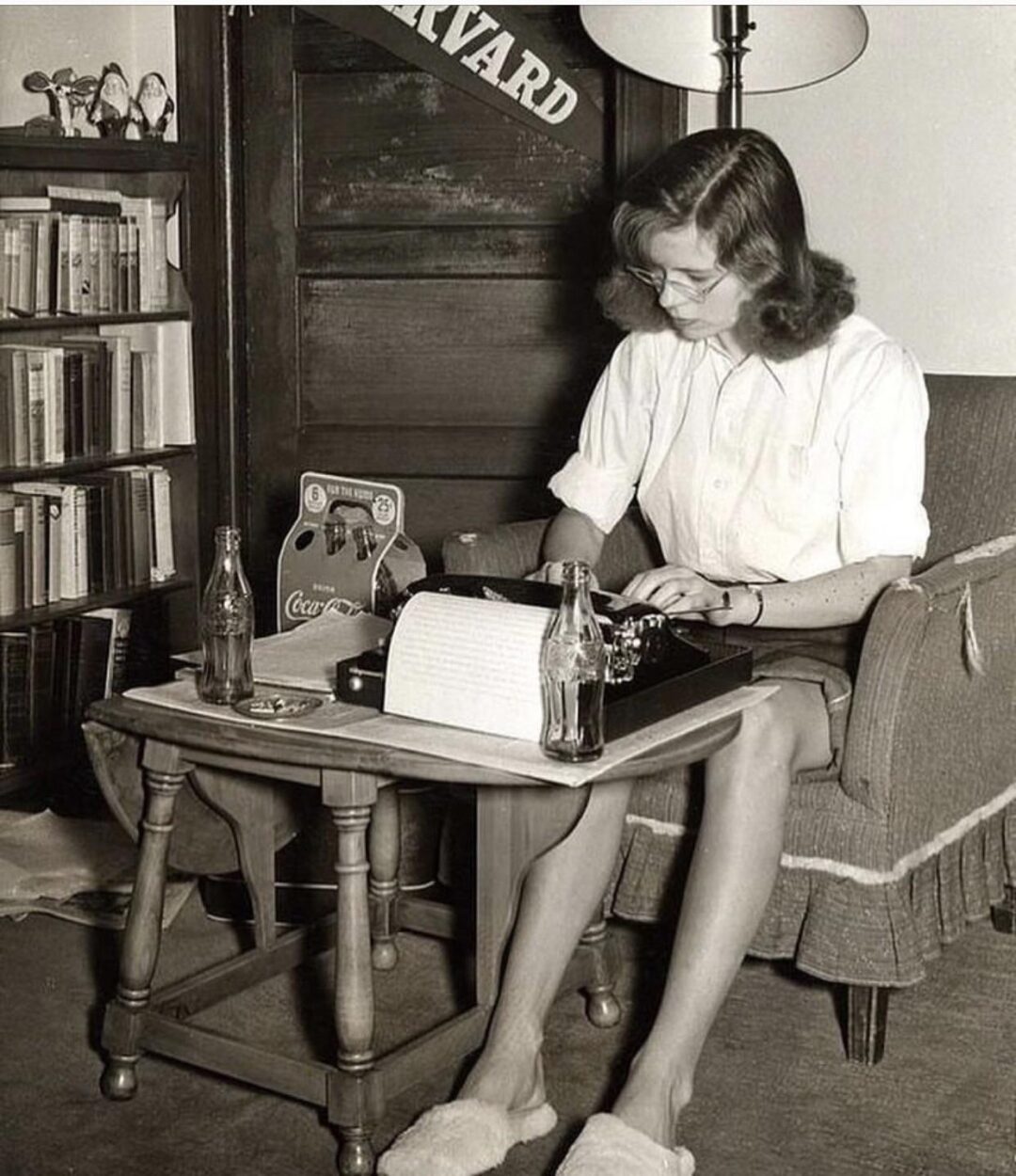 A woman in glasses and slippers types on a typewriter at a small table with Coca-Cola bottles, a lamp, bookshelves, and a "Harvard" banner in the background.