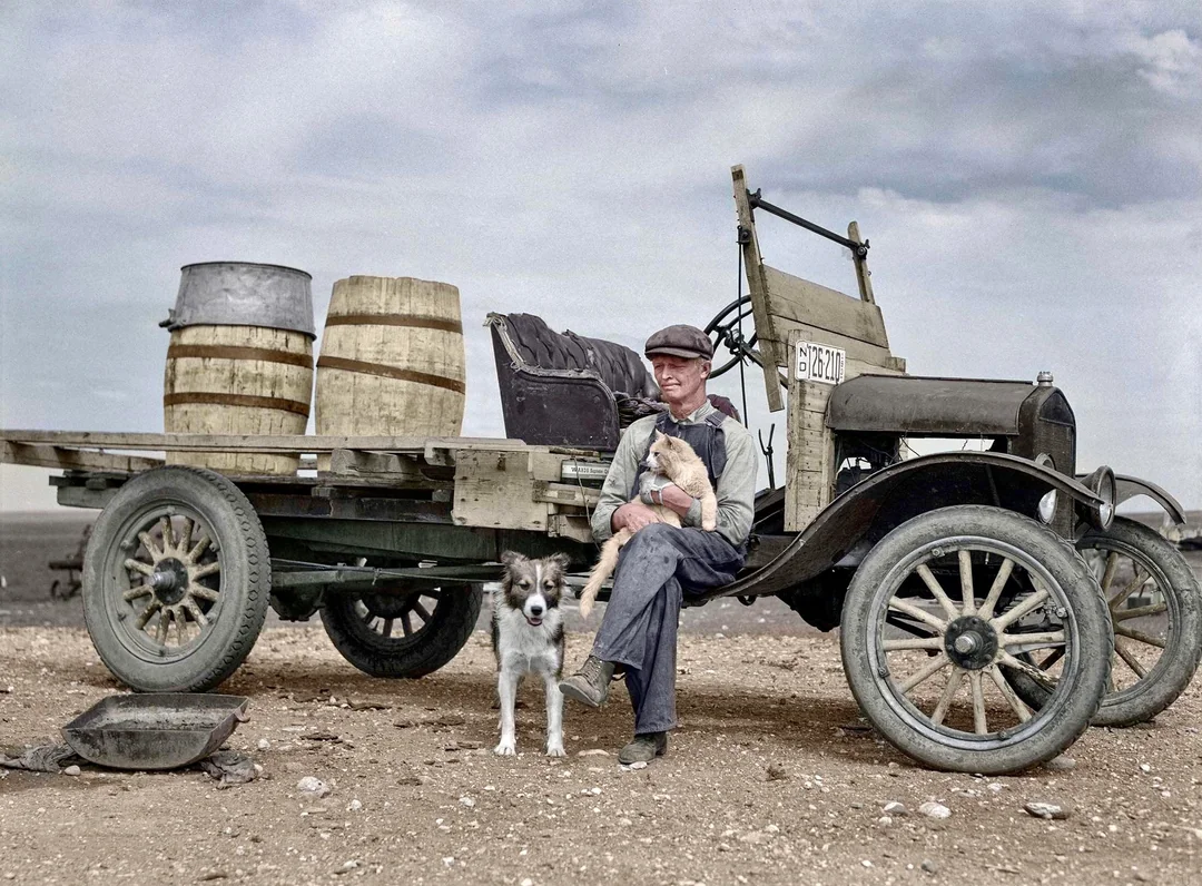 A man sits on the flatbed of an old truck with wooden barrels beside him, holding a child. A dog stands nearby on a rocky, barren landscape under a cloudy sky.