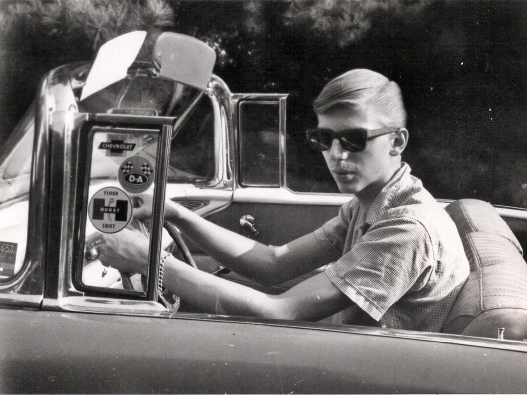 A young man wearing sunglasses and a short-sleeve shirt sits in the driver's seat of a vintage convertible car, holding the steering wheel. The car has a Chevrolet sticker on the window.