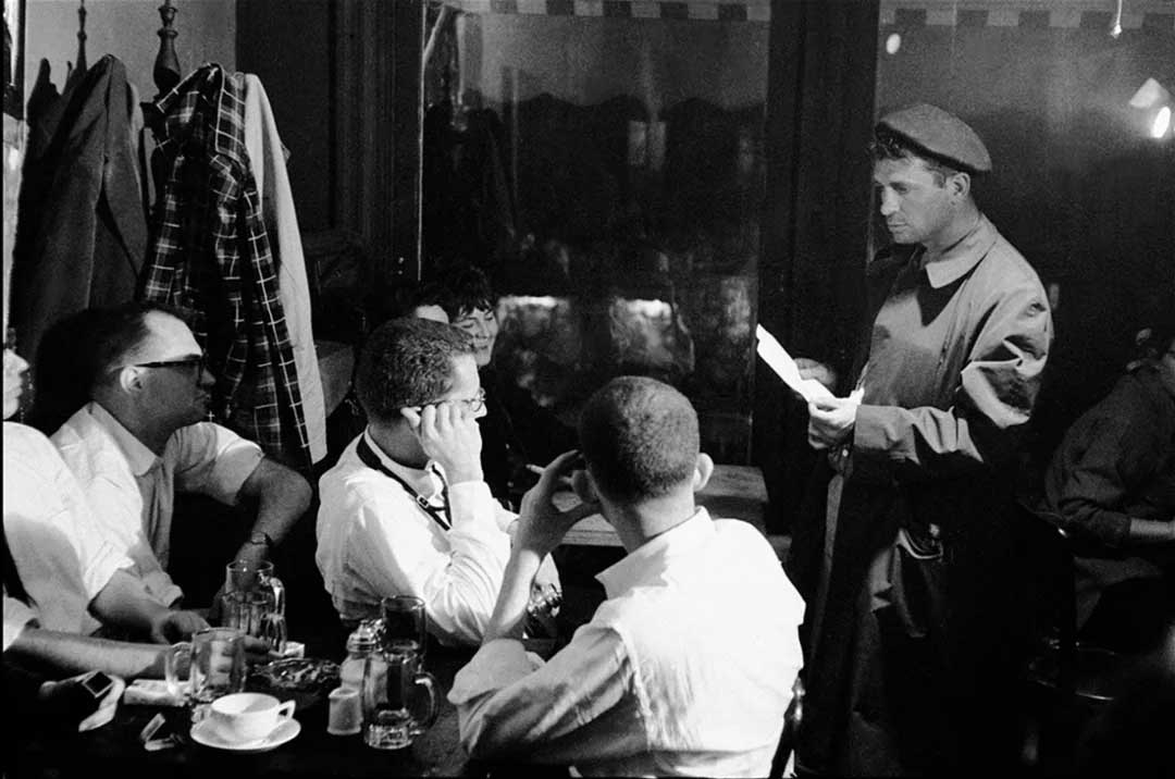 A man in a coat and cap stands in a dimly lit café, holding a paper, while four seated men listen attentively around a table with cups and dishes. Coats hang behind them, and a window reflects light in the background.