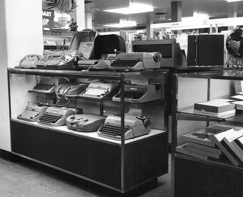 Black and white photo of a store display case featuring several vintage typewriters arranged on shelves, with books and globes visible in the background.