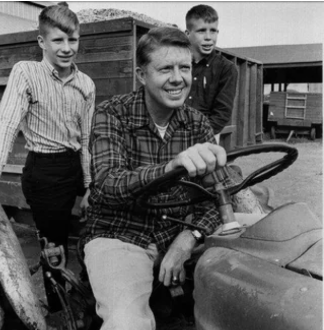 A man sits on a tractor smiling, with two boys standing beside him. All three wear casual, collared shirts. There are wooden trailers and a building in the background, suggesting a rural or farm setting.