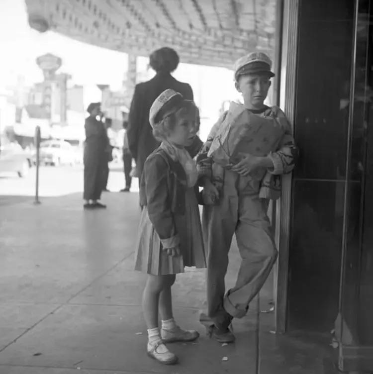 A young girl and boy, both wearing hats and holding hands, stand on a city sidewalk looking into a glass door. Other people and cars are visible in the background under a large marquee.