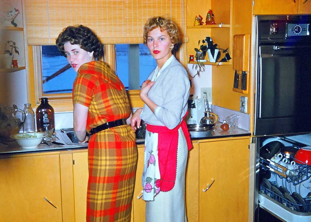 Two women in vintage clothing stand in a retro kitchen. One wears a red plaid dress, and the other a white outfit with a red apron. Both look toward the camera while standing near the sink and open dishwasher.