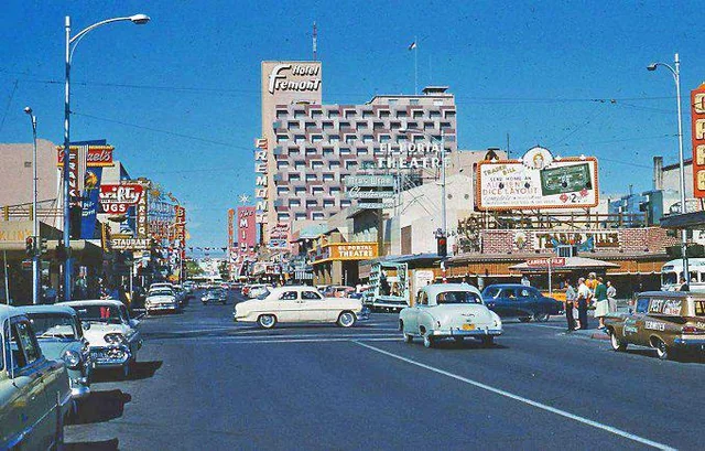 A busy city street scene from the mid-20th century with vintage cars, people crossing, and colorful storefronts and hotels, including the Fremont Hotel and a theater, under a bright blue sky.
