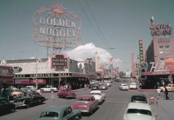 A busy 1950s street scene in Las Vegas with vintage cars, large neon signs for casinos like Golden Nugget and hotels, and people walking along the sidewalks under a bright sky.