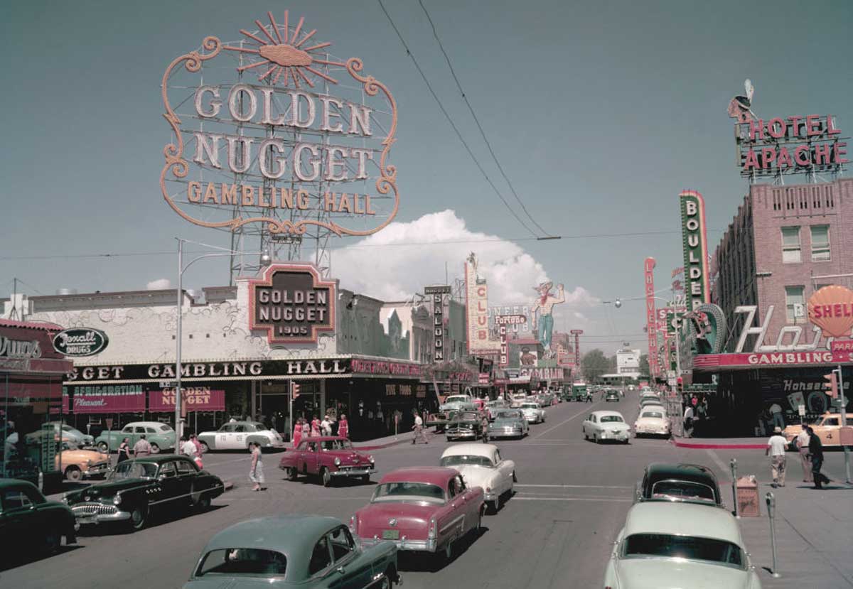 A busy 1950s street scene in Las Vegas with vintage cars, large neon signs for casinos like Golden Nugget and hotels, and people walking along the sidewalks under a bright sky.