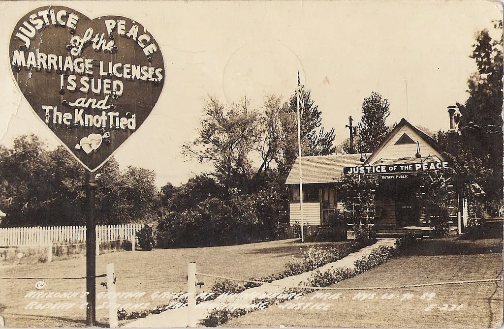 A vintage photo shows a building with a sign reading, “Justice of the Peace, Marriage Licenses Issued and The Knot Tied.” The building has a porch, flagpole, and surrounding trees and picket fence.