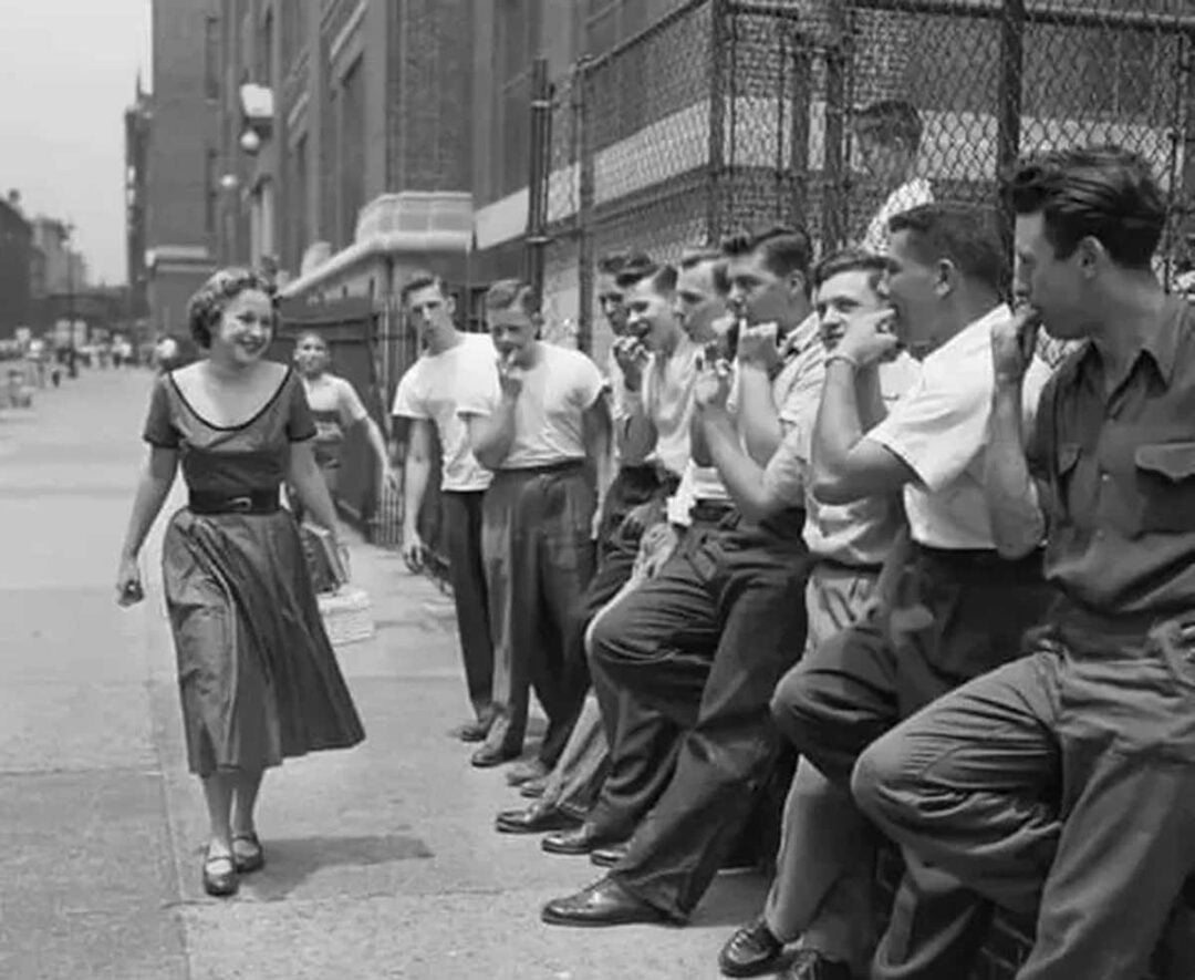 A woman in a dress walks past a group of young men leaning against a fence on a city street. The men watch her and appear to be whistling or making comments. The photo is black and white, suggesting it is from the mid-20th century.