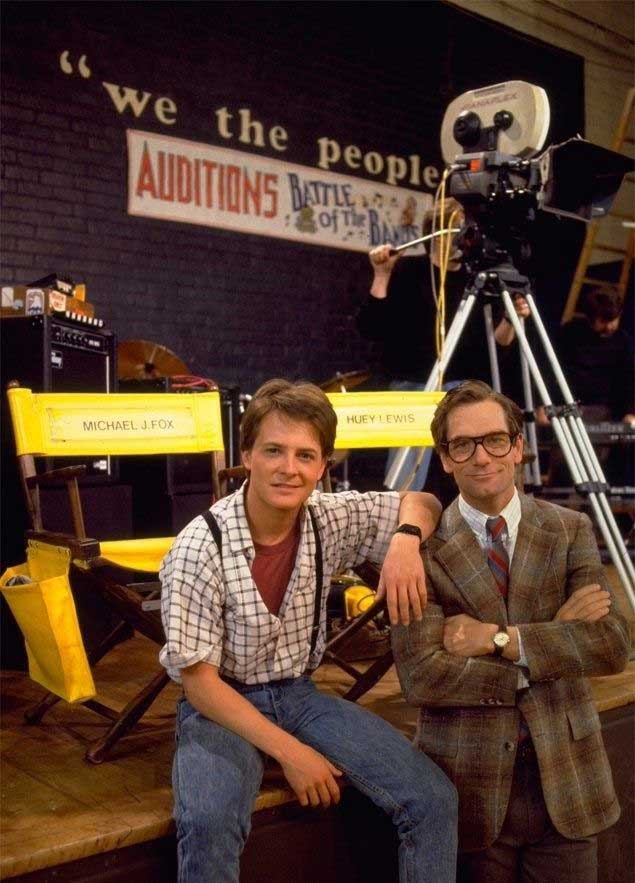 Two men pose on a film set in front of director’s chairs labeled “Michael J. Fox” and “Huey Lewis.” Behind them are a camera, a “Battle of the Bands Auditions” sign, and a partial “We the people” banner.