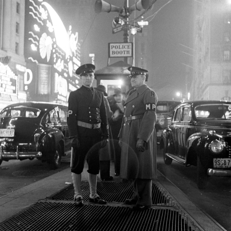 Two military police officers stand on a city street at night near a police booth, surrounded by vintage cars and bright neon signs. The scene appears to be from the 1940s.