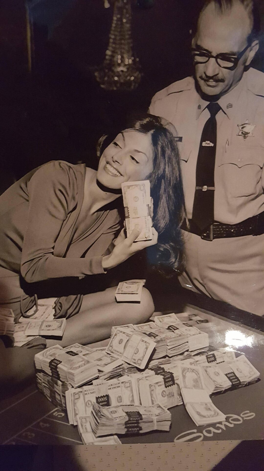 A woman smiling and holding a stack of cash sits beside a uniformed police officer. Numerous bundles of money are spread out on a table in front of them. The photo appears to be vintage and in black and white.