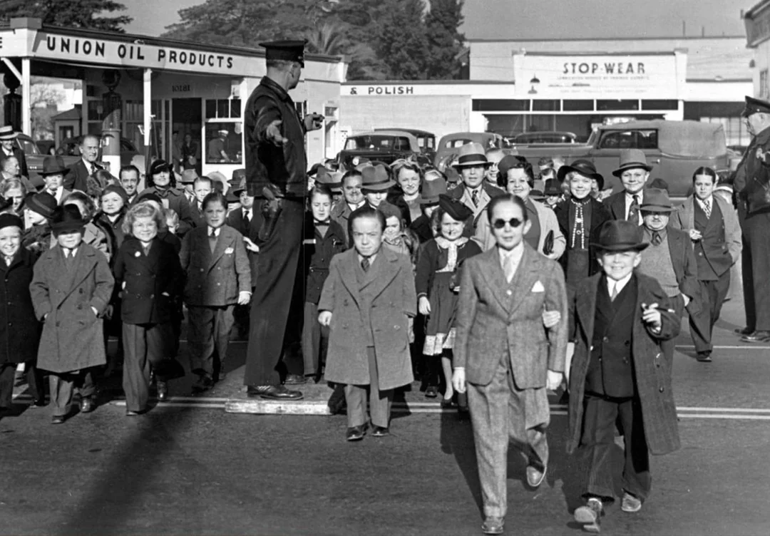 A group of children and adults, many in suits and hats, cross a street as a uniformed police officer stands on a platform directing traffic. Buildings and parked cars are visible in the background.